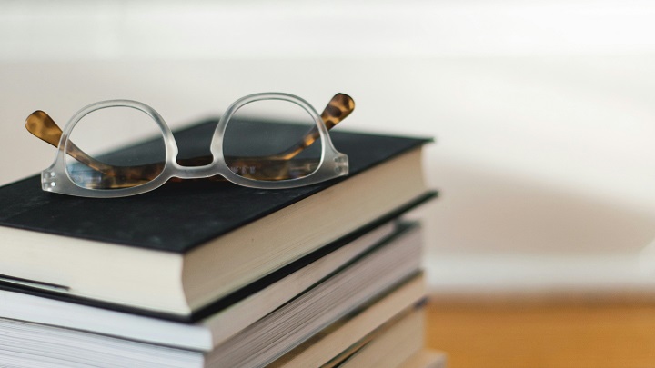 Stos książek z okularami: okulary z przezroczystymi oprawkami i brązowymi szkłami leżą na czarnej okładce najwyższej książki.