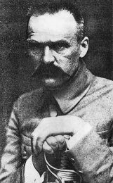Józef Piłsudzki ubrany w mundur. W rękach trzyma szablę, którą się podpiera.